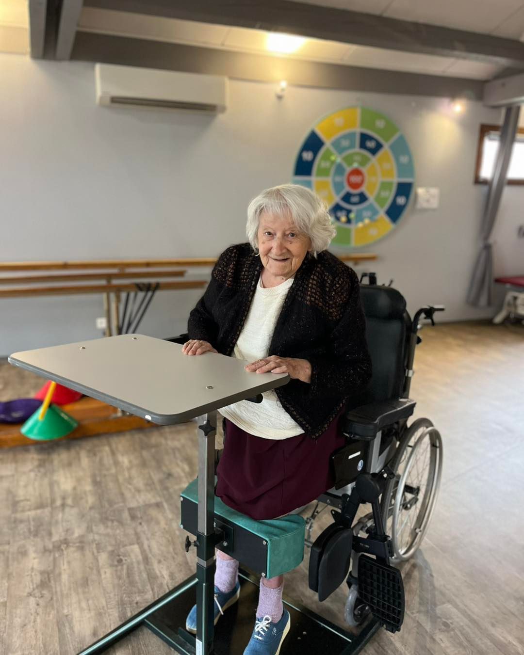 Une femme souriante en fauteuil roulant tient une table dans une salle d'activités intérieure lumineuse