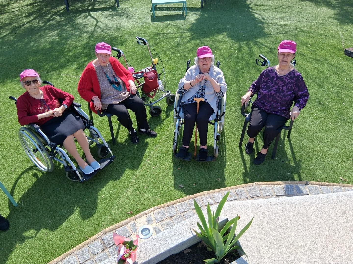 Quatre personnes coiffées de chapeaux roses sont assises en fauteuil roulant à l'extérieur, sur une pelouse.