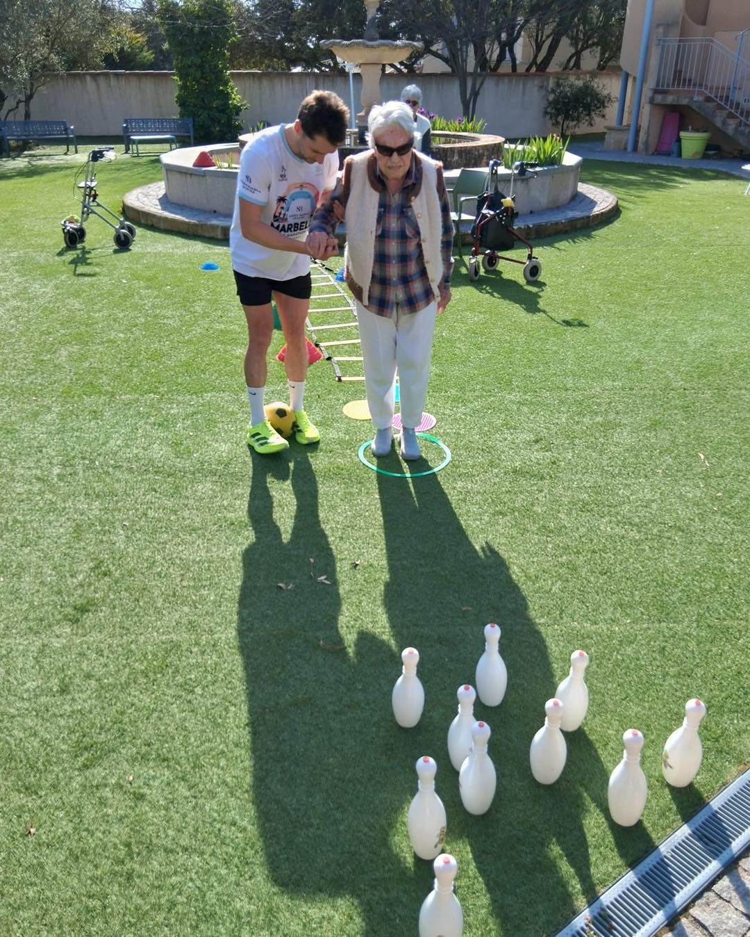 Deux personnes jouent aux boules sur une pelouse ensoleillée, à côté d'une fontaine.