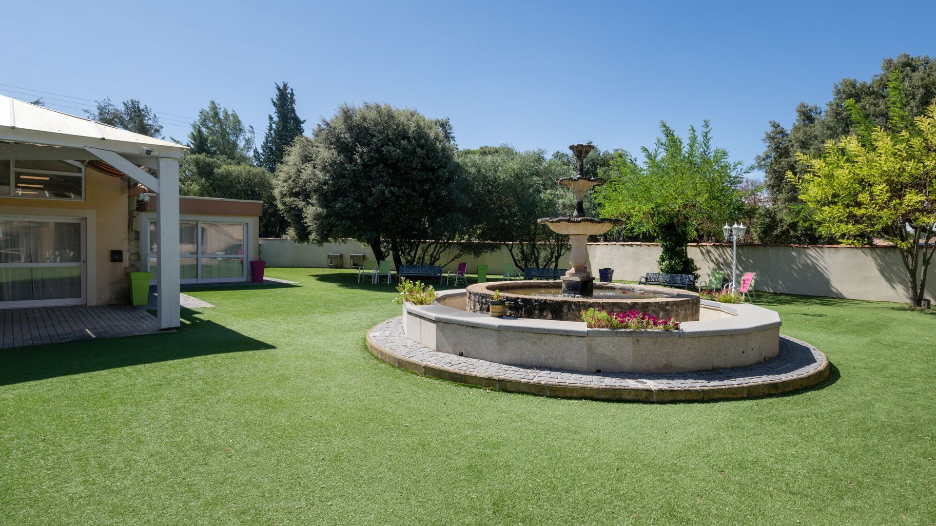 Un jardin agrémenté d'une fontaine en pierre, de pelouse et d'un bâtiment aux couleurs claires.