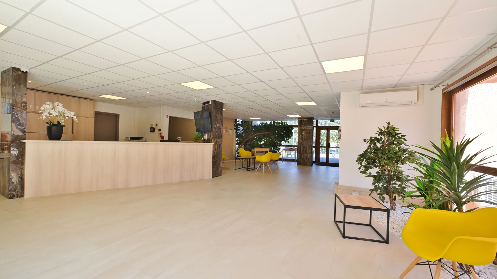 Un hall lumineux avec un long comptoir de réception, des fauteuils jaunes et des plantes d'intérieur.