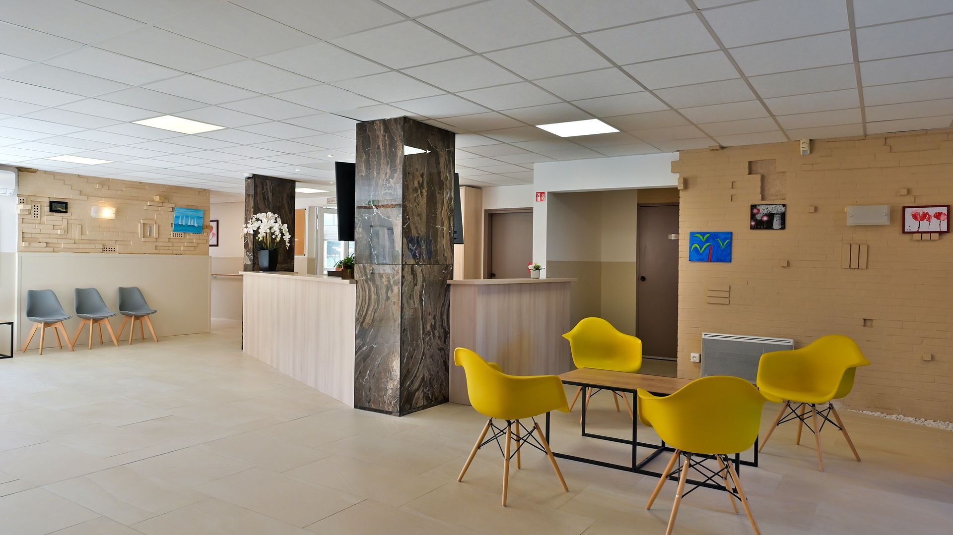 Une salle d'attente aux murs clairs, un comptoir d'accueil, un pilier central en pierre et des fauteuils modernes jaune vif.