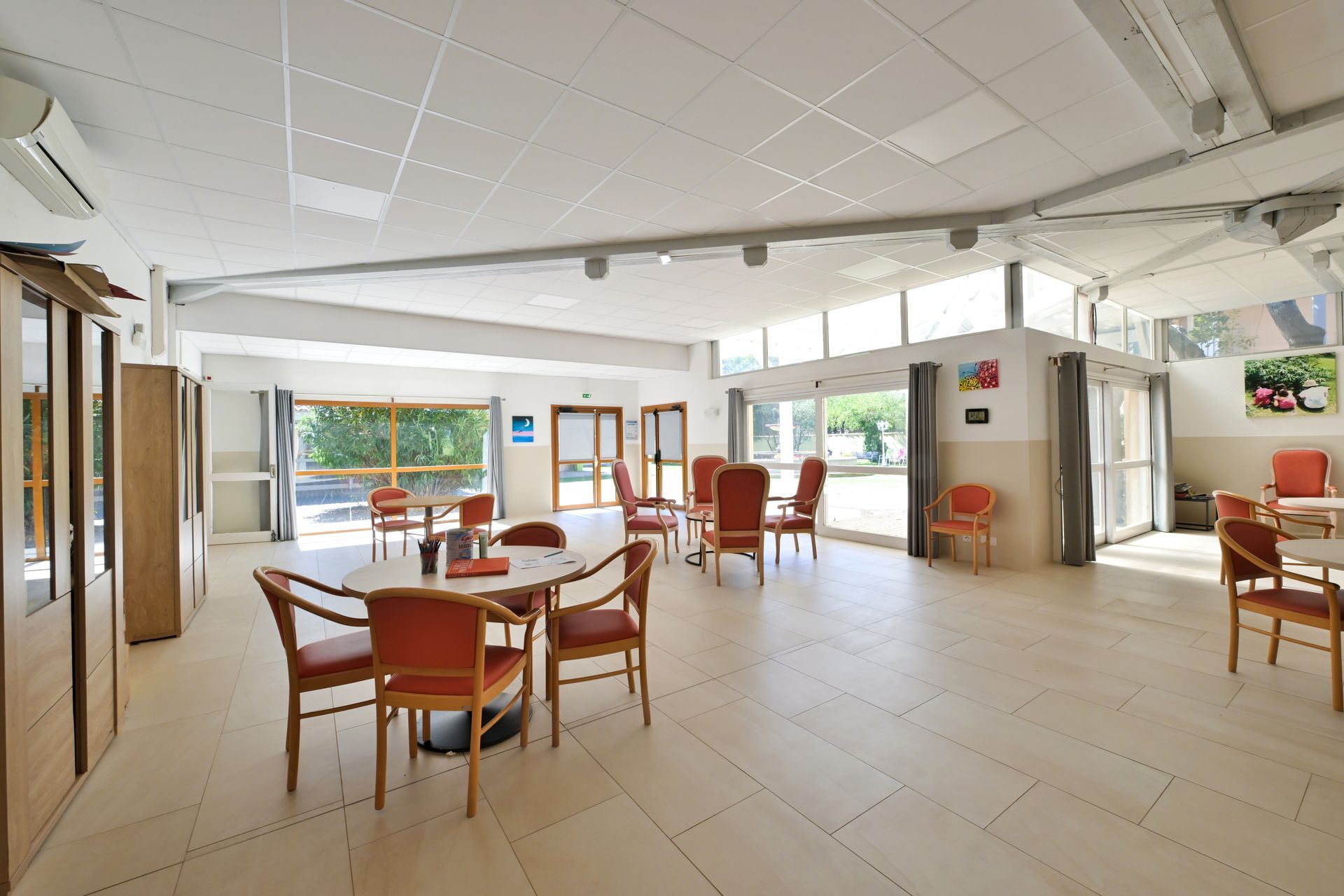 Une salle à manger spacieuse et lumineuse avec un sol carrelé, plusieurs tables rondes et des chaises aux coussins orange.
