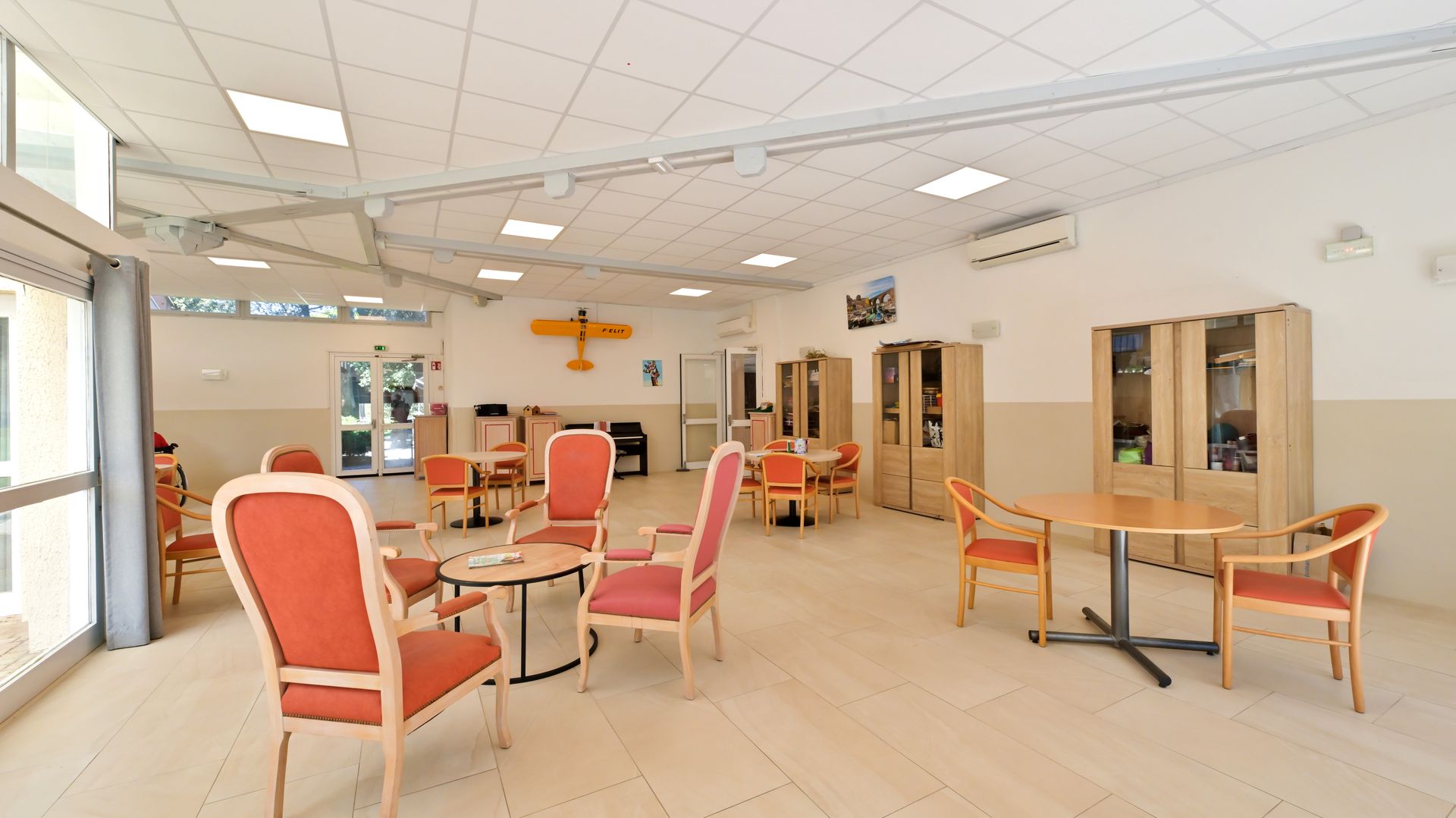 Une salle commune lumineuse et spacieuse avec un sol carrelé, des tables et des chaises à coussins rouges.
