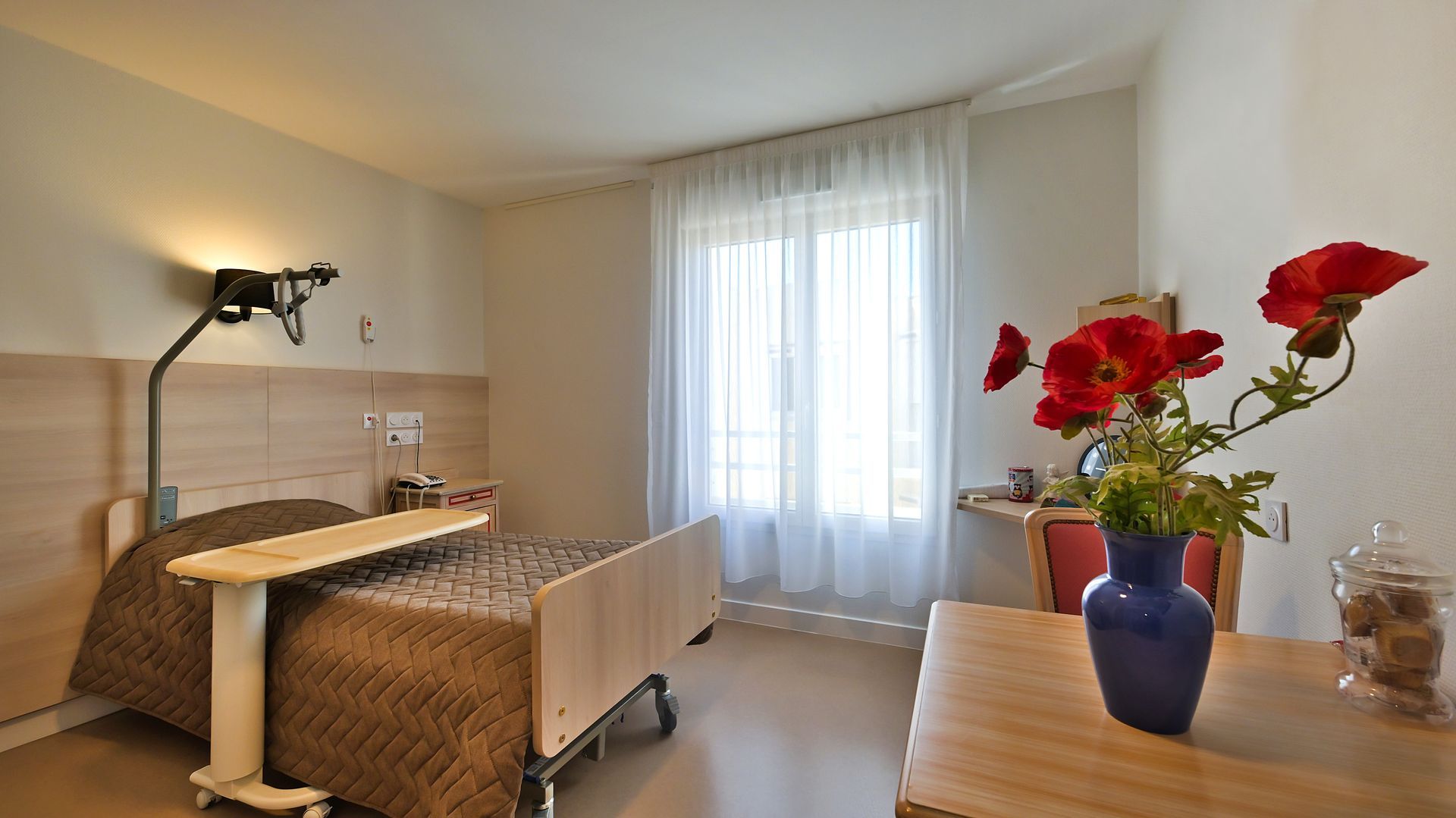 Une chambre d'EHPAD avec un lit, une table, une fenêtre et un vase de fleurs rouges sur un bureau.