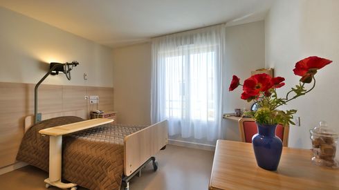 Une chambre d'EHPAD avec un lit réglable, une table de chevet et une fenêtre avec des rideaux.