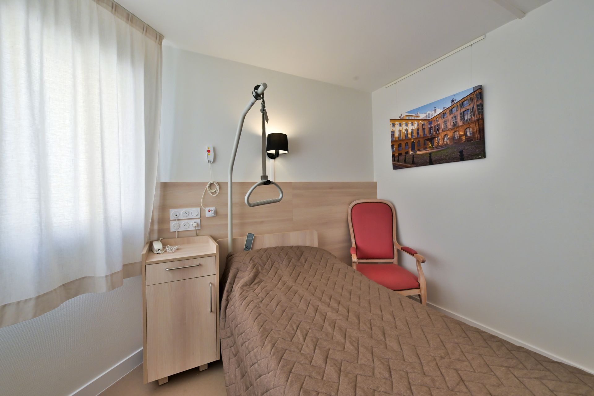 Une chambre d'EHPAD avec un lit simple, un lève-personne, une table de chevet, une chaise rouge et un tableau au mur.