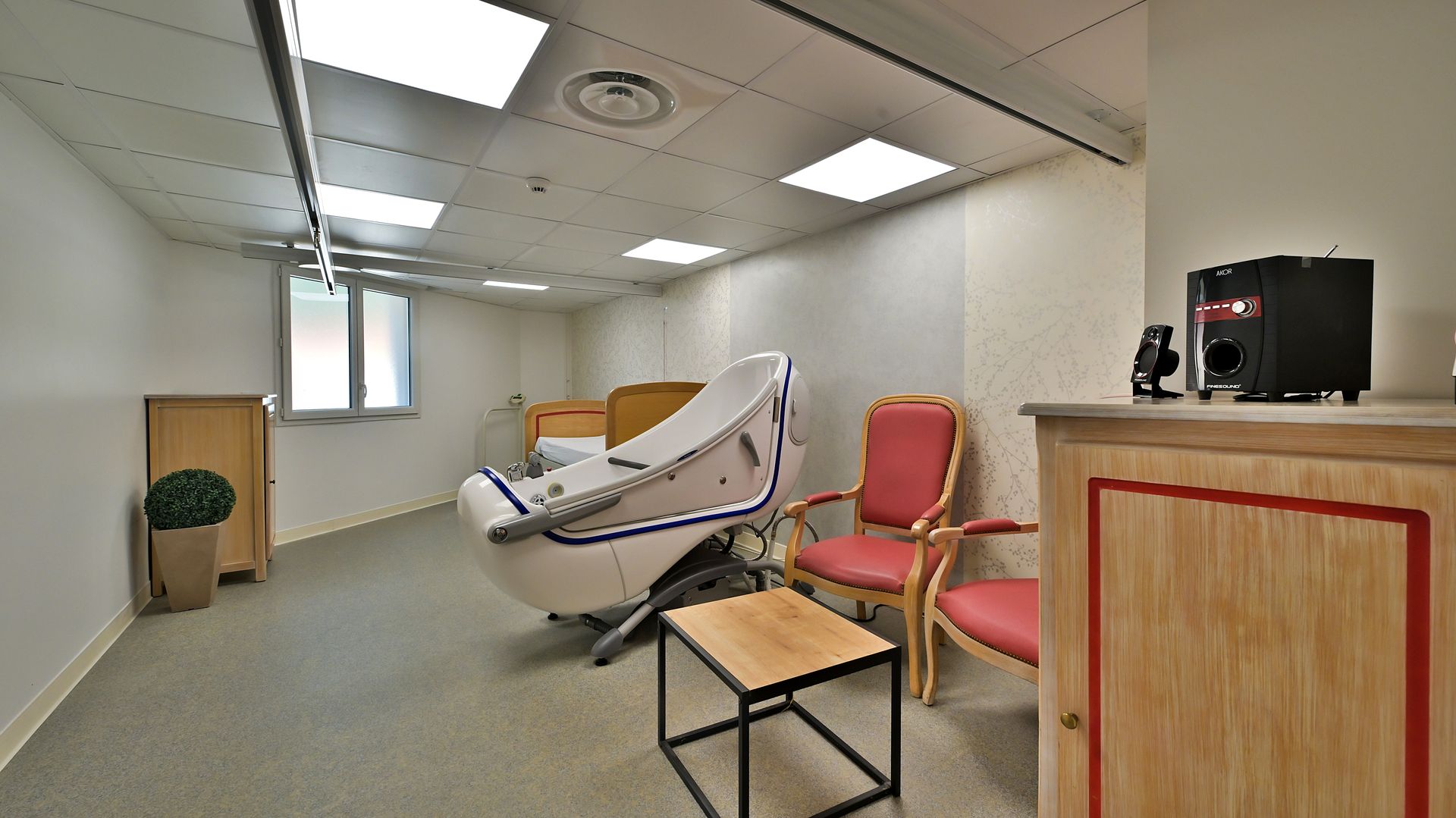 Une pièce avec une baignoire médicale, deux chaises, une table d'appoint et une armoire en bois.