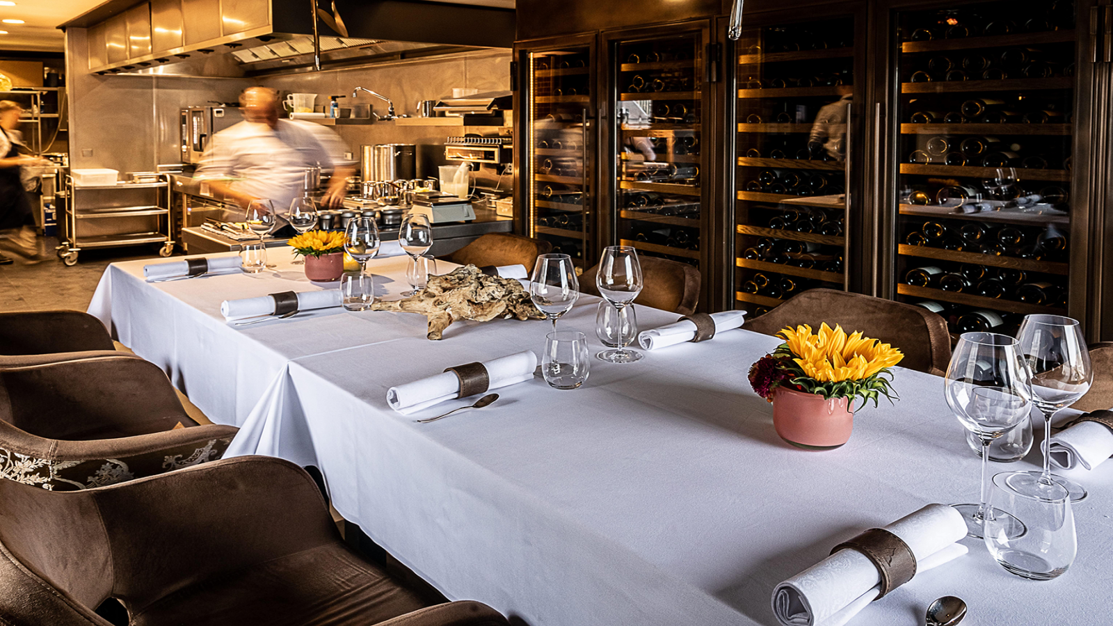Lange eettafel gedekt Chefs Table voor een maaltijd in een restaurant, met een chef-kok zichtbaar op de achtergrond.