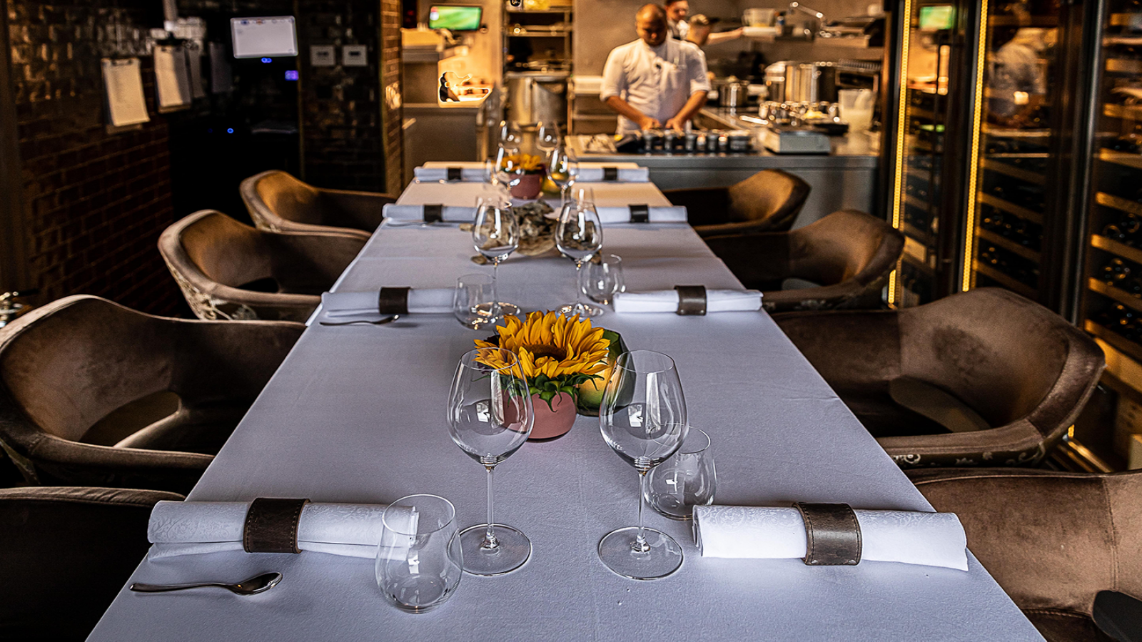Lange eettafel Chef's Table met elegant glaswerk en een bloemstuk; chef-kok op de achtergrond.