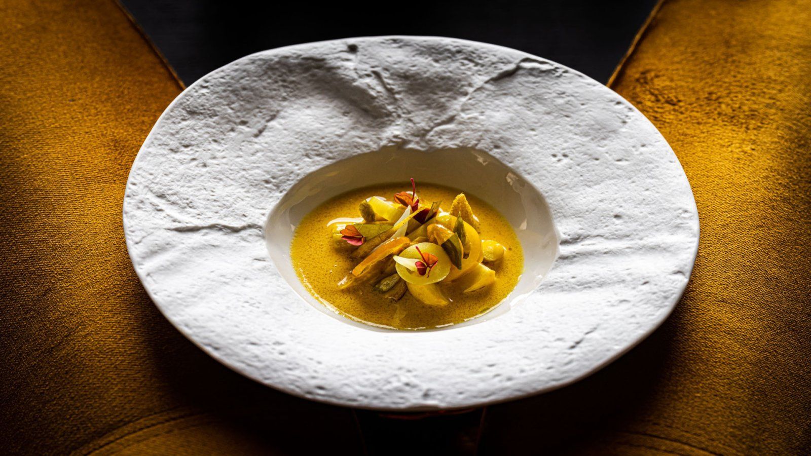 restaurant Bougainville: Een witte, getextureerde kom met een gele bouillon met groenten, op een gouden ondergrond.