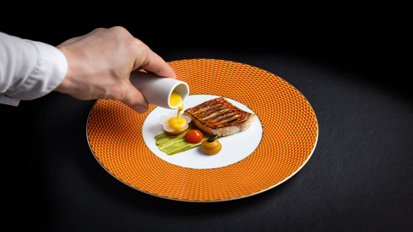 restaurant Bougainville De chef giet saus over gegrilde vis op een oranje bord met groenten. Zwarte achtergrond.