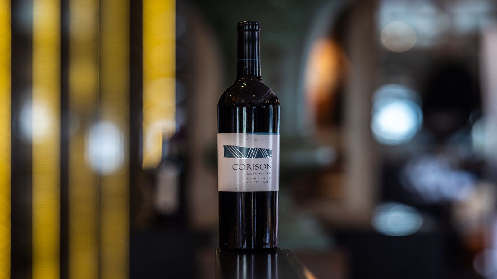 Een fles rode wijn met een wit etiket, tegen een onscherpe achtergrond met gele en zwarte verticale strepen.