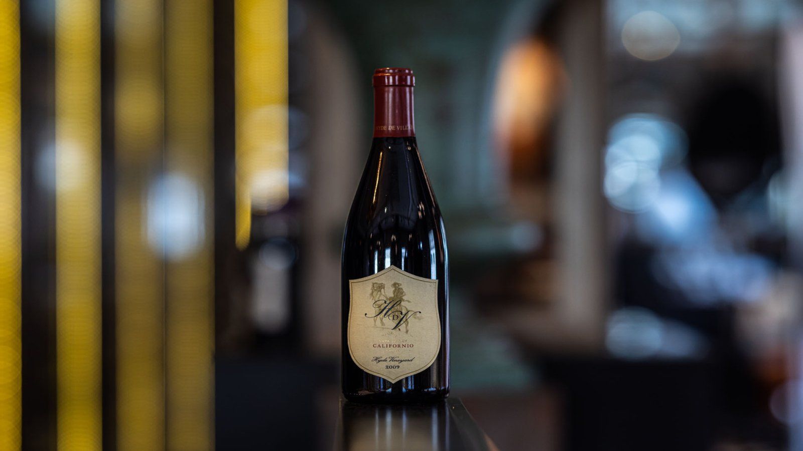 Een fles rode wijn op een bar, gouden en zwarte strepen op de achtergrond, een persoon is vaag te zien op de achtergrond.