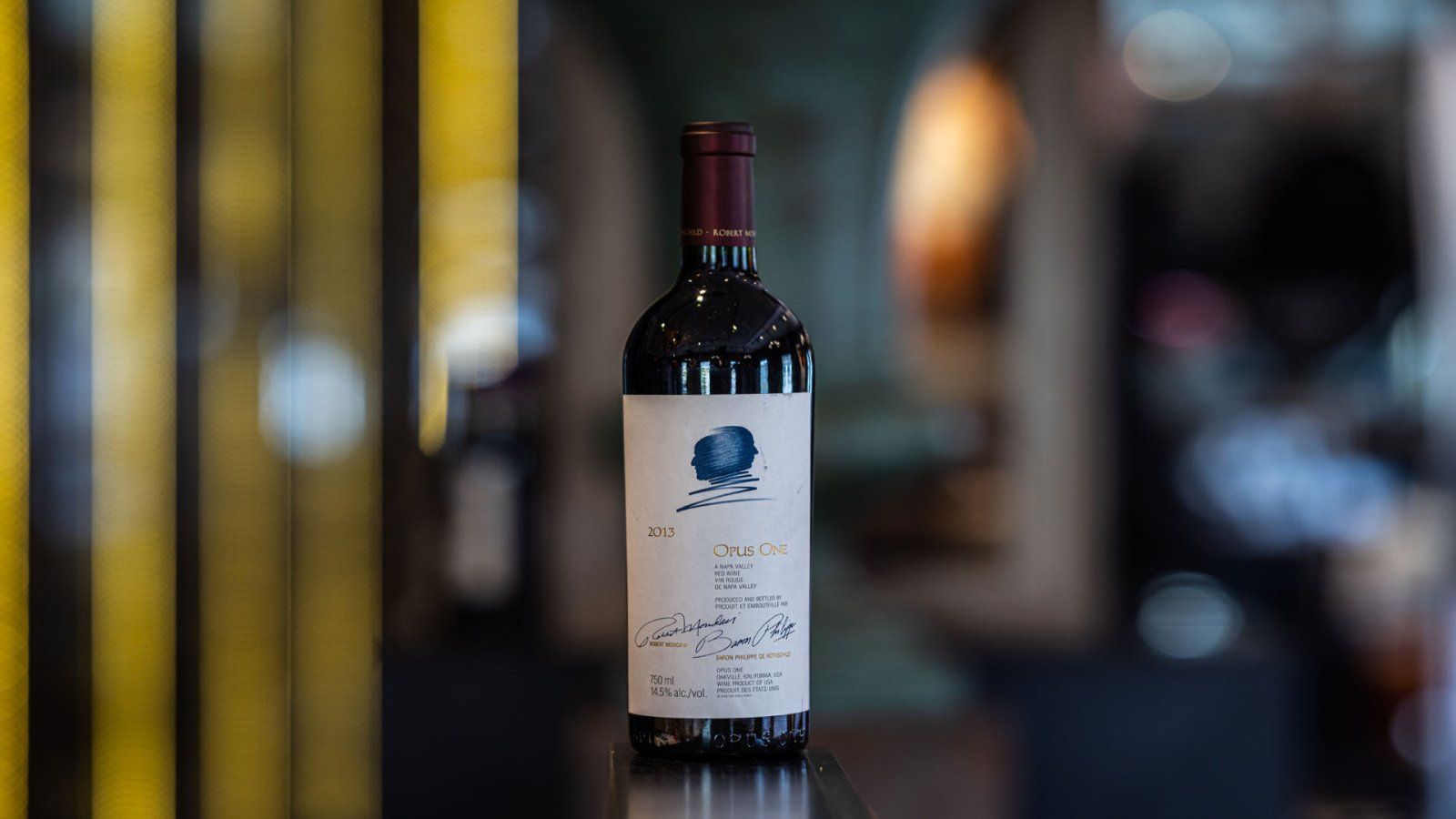 Een fles rode wijn met een blauw logo op een reflecterend zwart oppervlak in een slecht verlichte kamer.