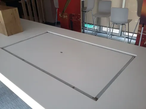 Mesa blanca con abertura rectangular empotrada; sillas y vista parcial de un fondo rojo.