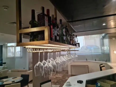 Bar con botellas de vino en un estante y copas colgando debajo. Fotografía interior con barra y asientos.