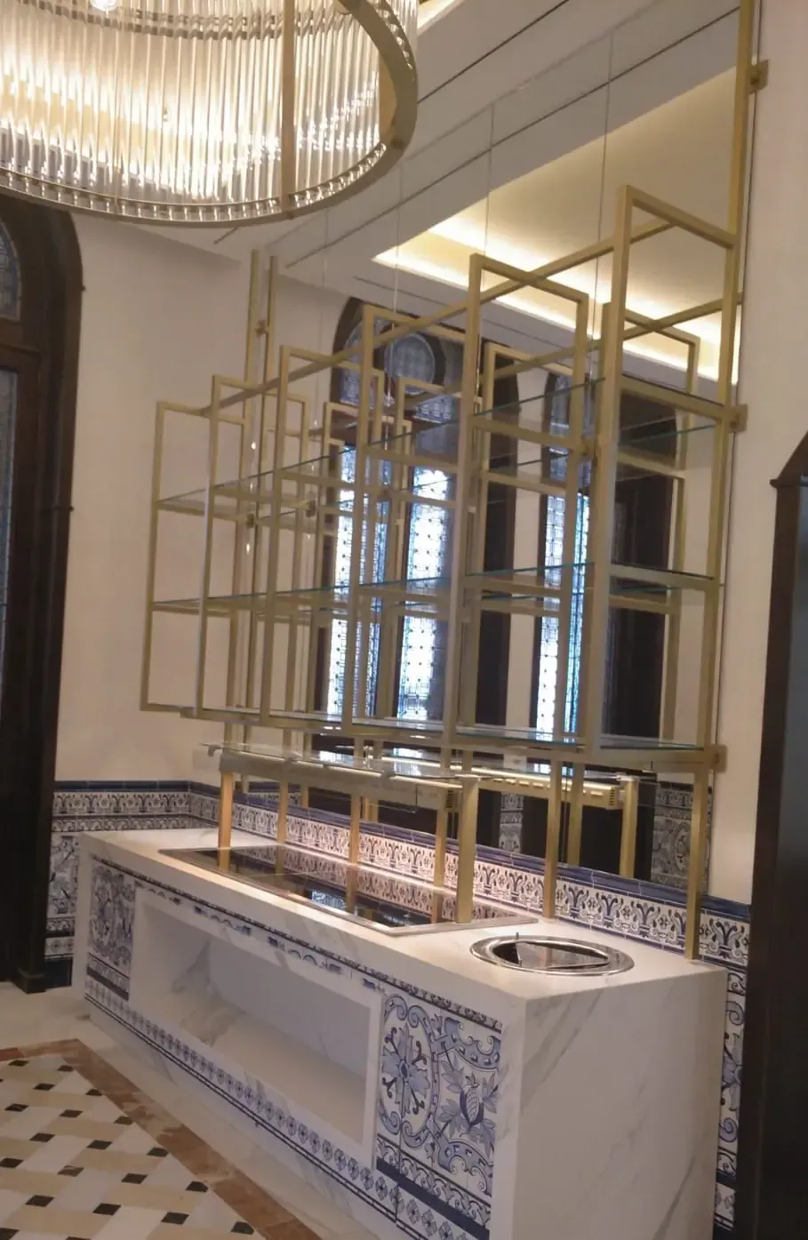 Estantería de metal dorado sobre una encimera de mármol con azulejos azules. Pared con espejos y lámpara de araña al fondo.