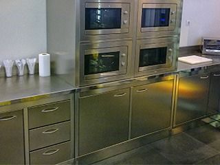 Cocina comercial de acero inoxidable con cajones, gabinetes y cuatro microondas incorporados.