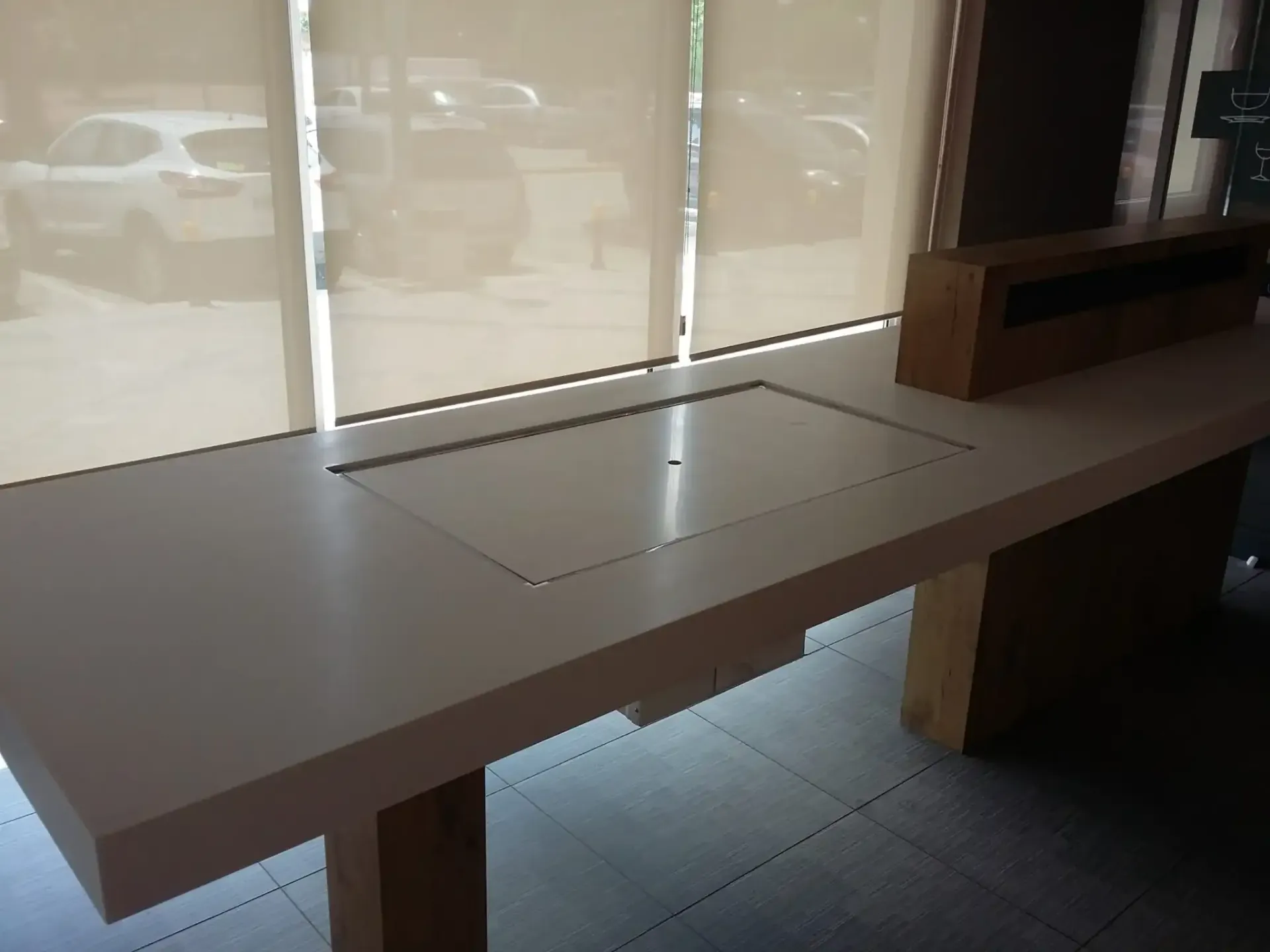 Un largo mostrador blanco con un panel empotrado, cerca de una ventana con cortinas; autos visibles afuera.