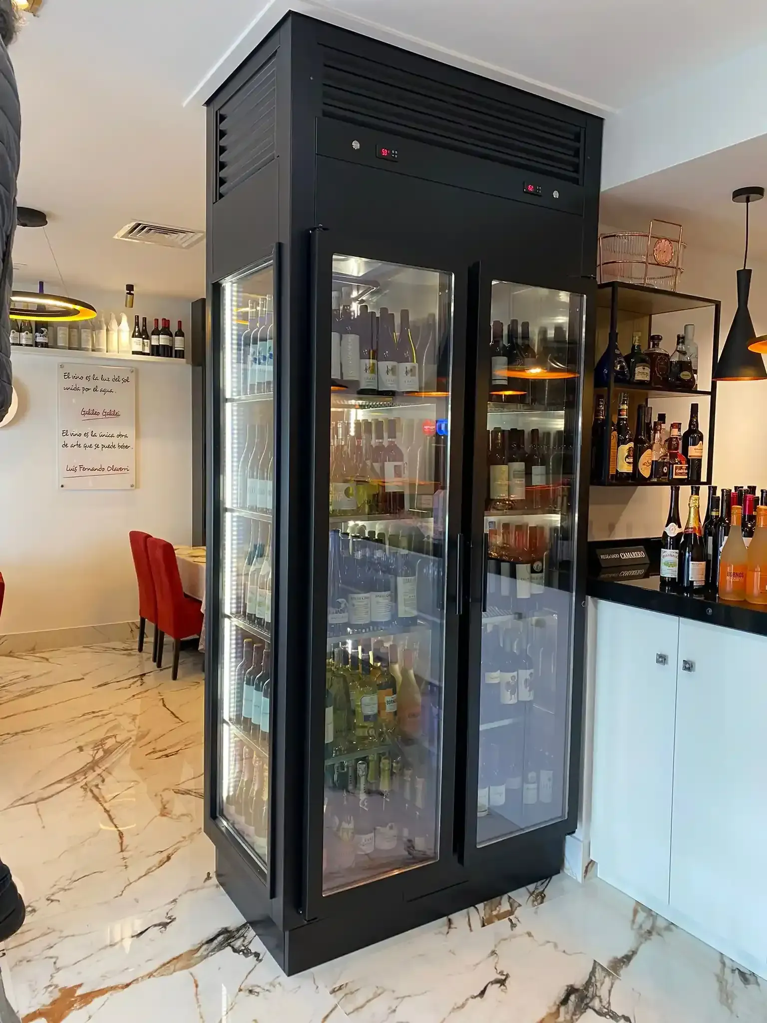 Enfriador de vino negro con puertas de vidrio que exhibe botellas de vino en un restaurante.