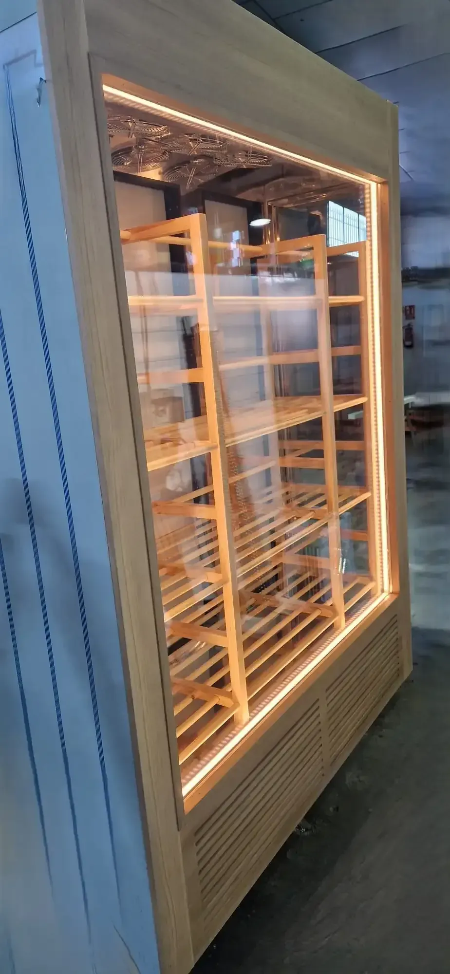 Botellero de madera con frontal de cristal, iluminado con luces cálidas, en el interior de un almacén.