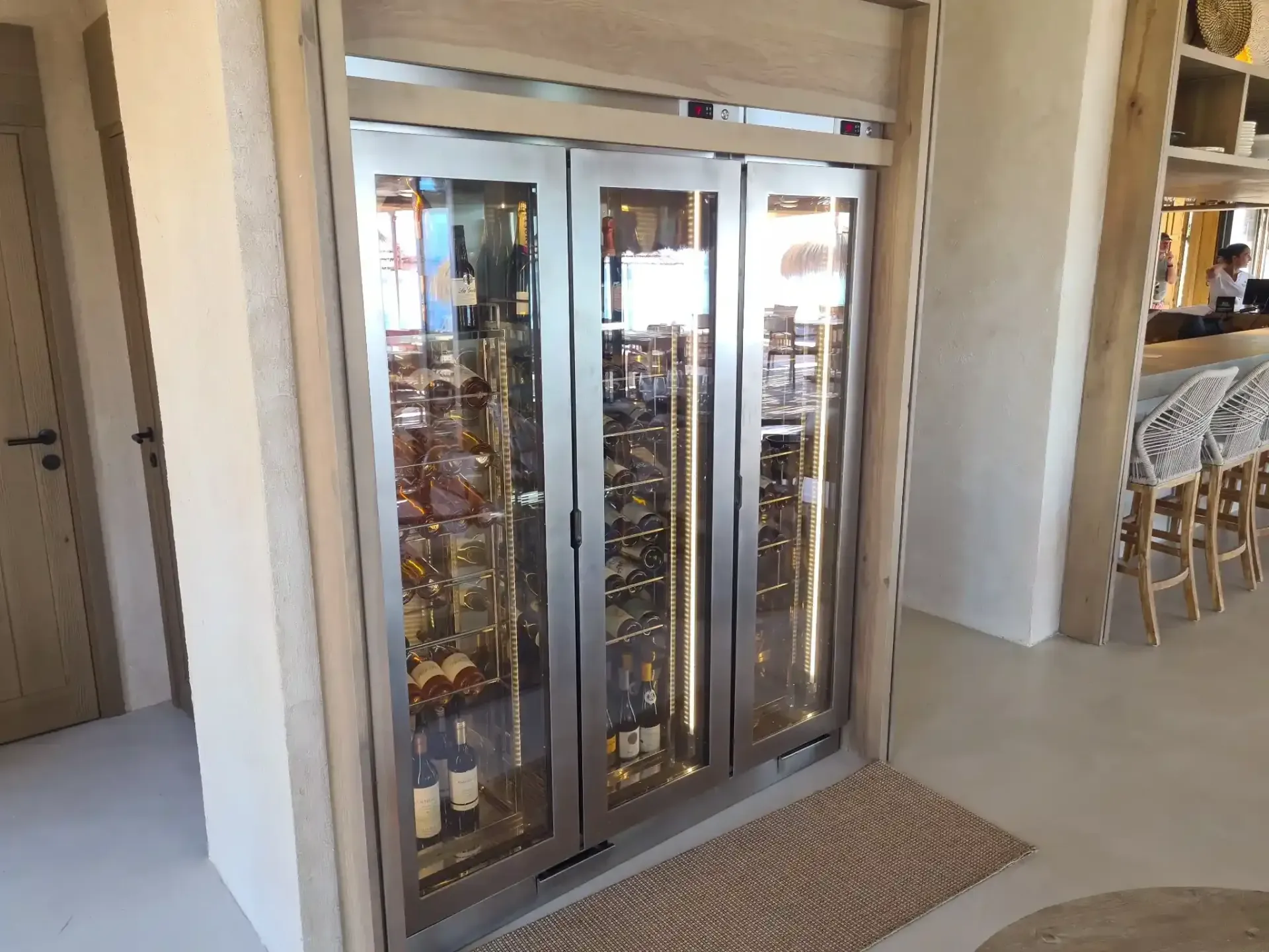 Bodega con puertas de cristal situada en un marco de madera clara, llena de botellas.