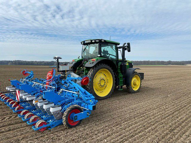 Ein grüner Traktor zieht eine blau-rote Sämaschine über ein gepflügtes Feld unter einem bewölkten Himmel