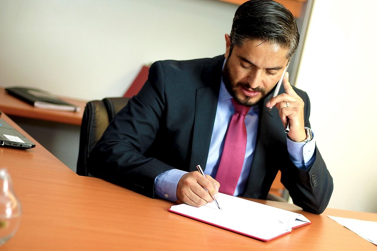 Homem de terno, ao telefone, escrevendo em um papel na mesa do escritório.