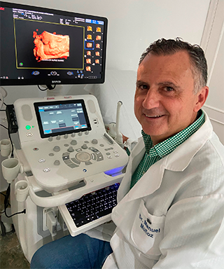 Médico sonriendo junto a un ecógrafo, con la pantalla mostrando la imagen. Lleva bata y camisa verde.