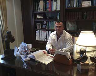 Un hombre con bata blanca se sienta en un escritorio con libros y una lámpara, con estanterías al fondo.