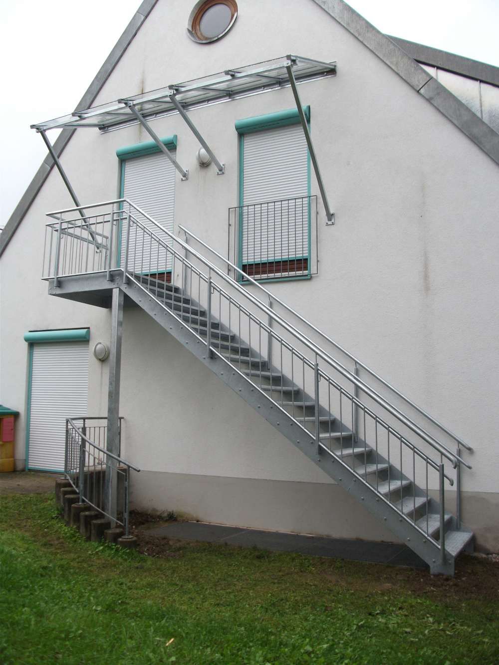 Außenrampe mit Metallhandlauf entlang eines gefliesten Gehwegs neben einer weißen Mauer und Bäumen
