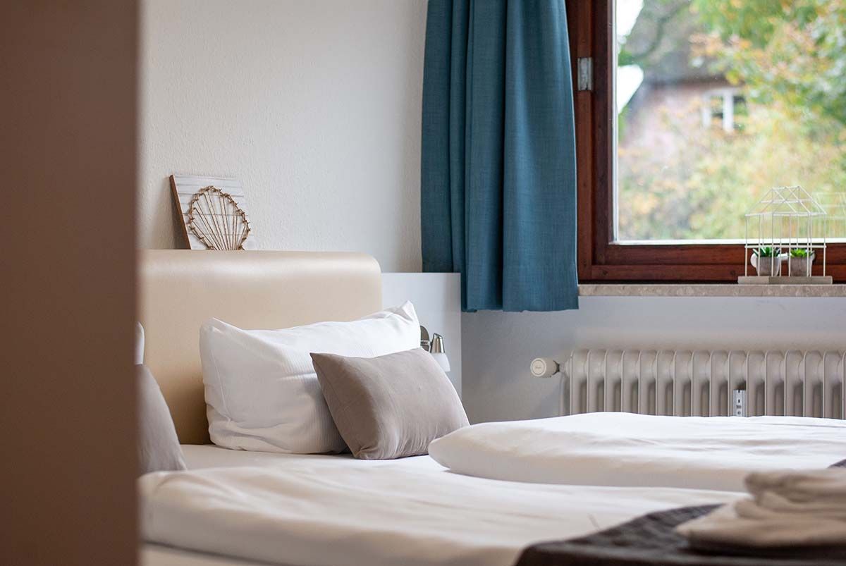 Gemütliches Schlafzimmer mit ordentlich gemachtem Bett, weißen Kissen, türkisfarbenen Vorhängen und einem Fenster mit einem Heizkörper darunter.