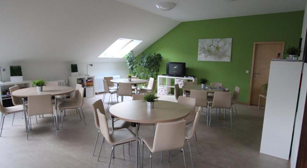Helle Cafeteria mit runden Tischen, beigefarbenen Stühlen, grüner Akzentwand, Oberlicht und Topfpflanzen.