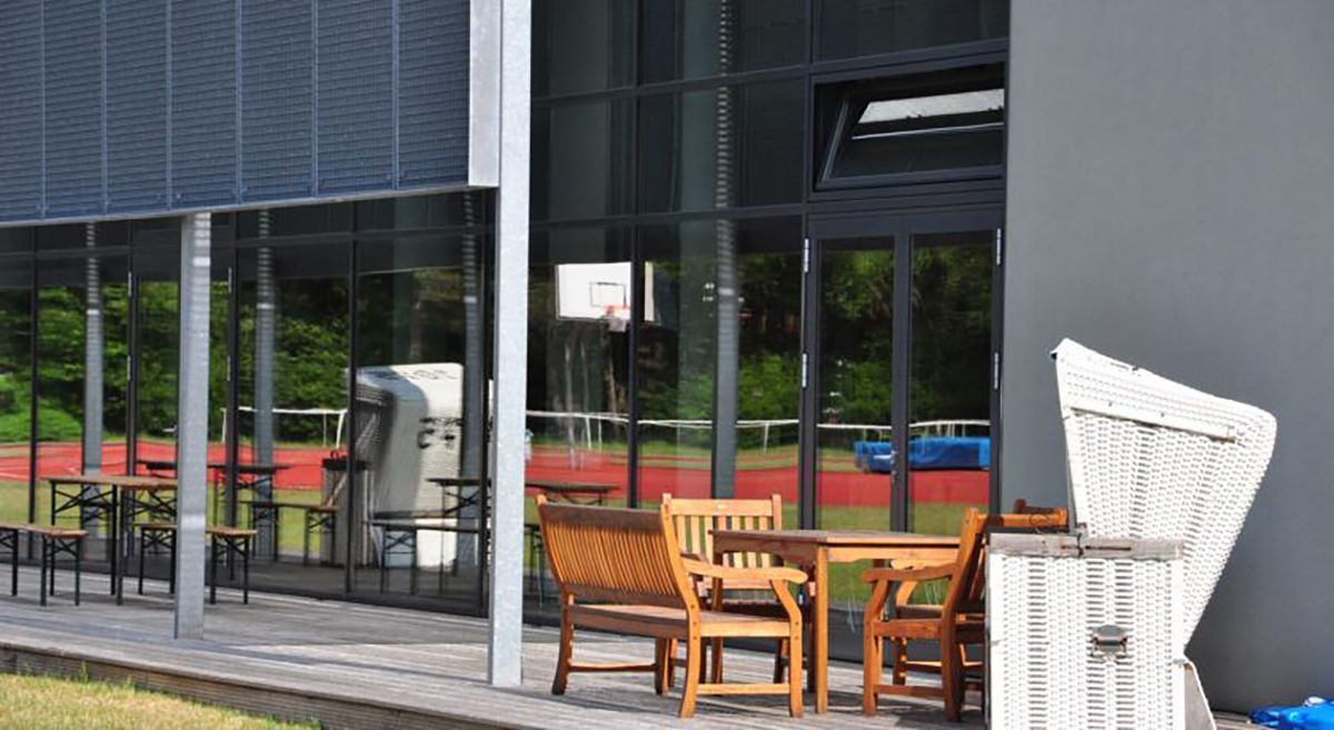 Terrasse mit Korbstühlen und Holztisch neben einem modernen Gebäude mit großen Glasfenstern und Jalousien.