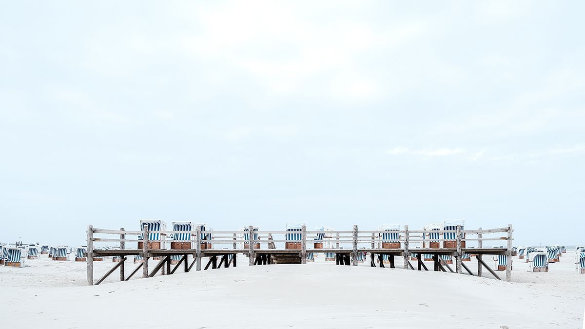 Breiter, weißer Sandstrand mit einem hölzernen Rettungsschwimmerstand und Reihen von Strandstühlen unter einem hellen Himmel