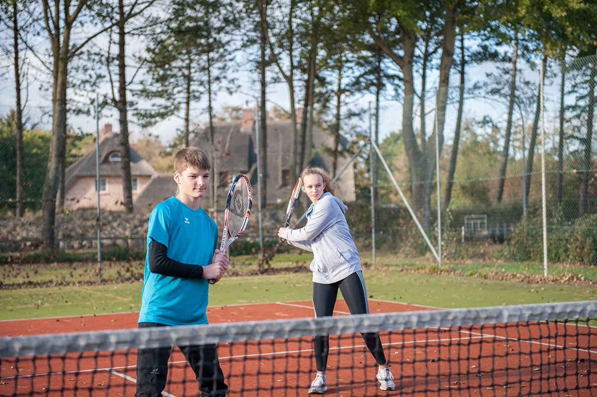 Zwei Personen spielen Tennis auf einem Freiluftplatz; im Vordergrund ist ein Netz zu sehen, im Hintergrund Bäume.