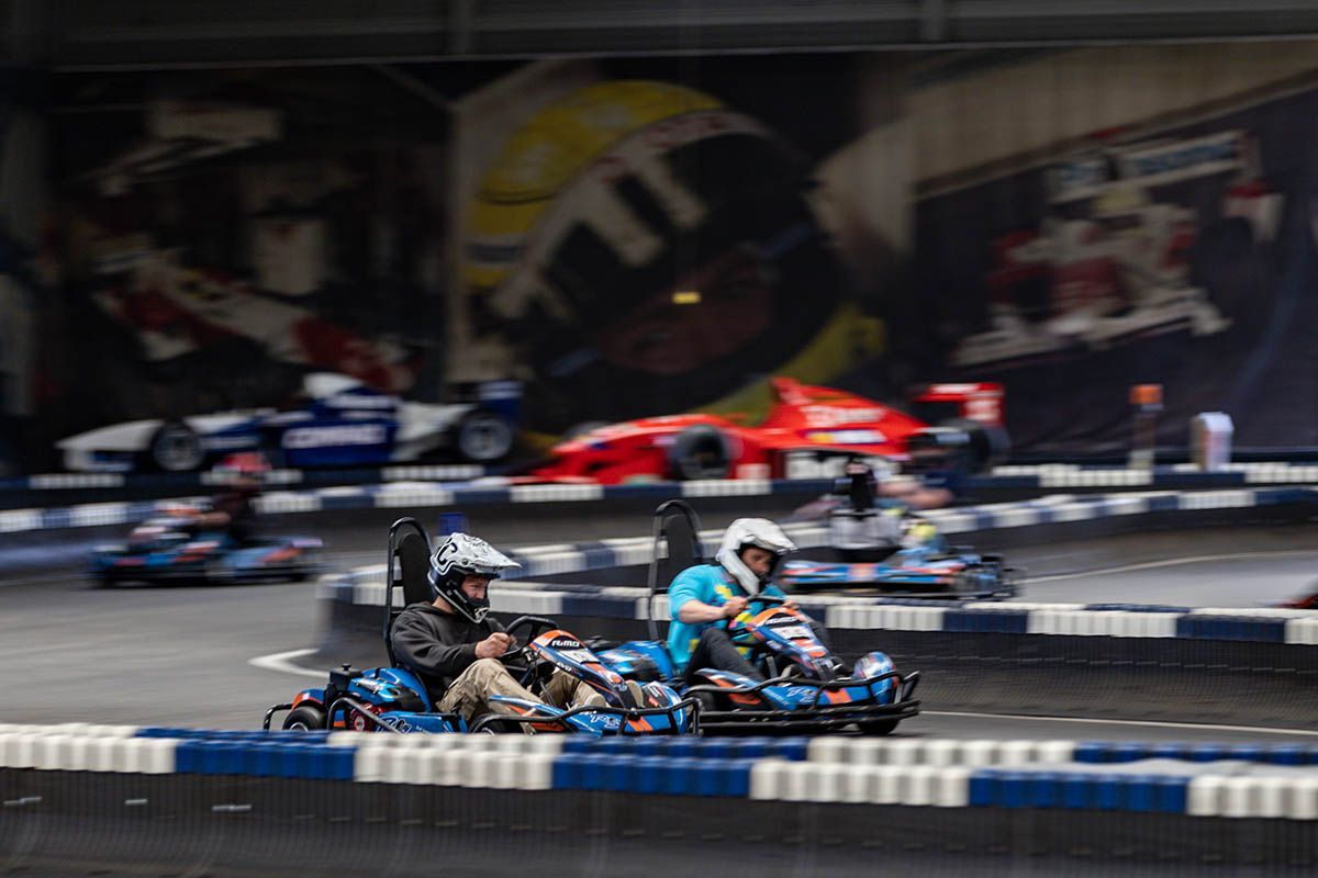 Zwei Go-Karts rasen nebeneinander auf einer Indoor-Rennstrecke; im Hintergrund sind Fahrer und Zuschauer verschwommen zu erkennen.