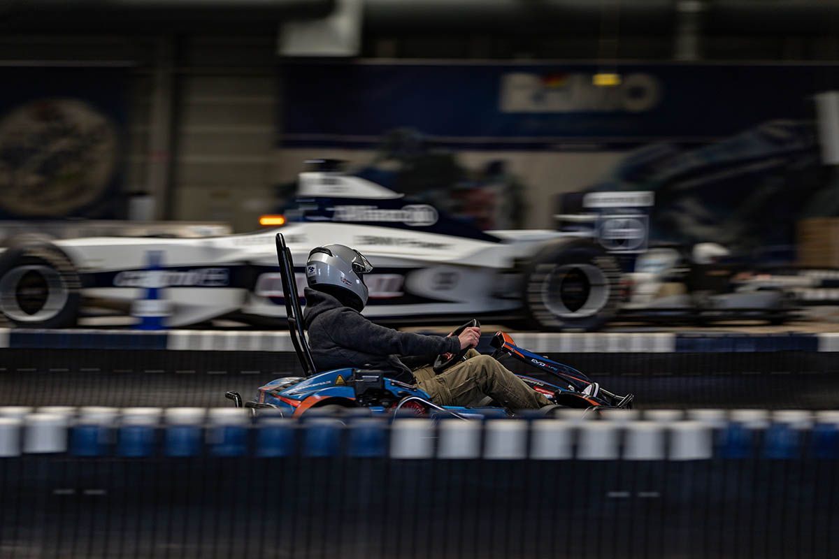 Verschwommener Go-Kart-Fahrer rast auf einer Rennstrecke; er trägt einen Helm und einen dunklen Anzug; im Vordergrund sind Barrieren zu sehen.