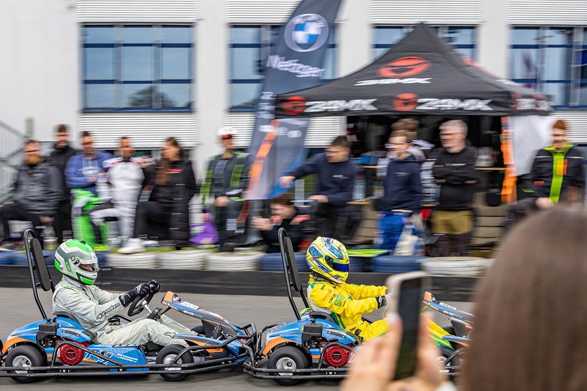 Zwei Kartrennfahrer in silber-blauen Karts, einer mit grünem, der andere mit gelbem Helm, rasen an den Zuschauern vorbei.