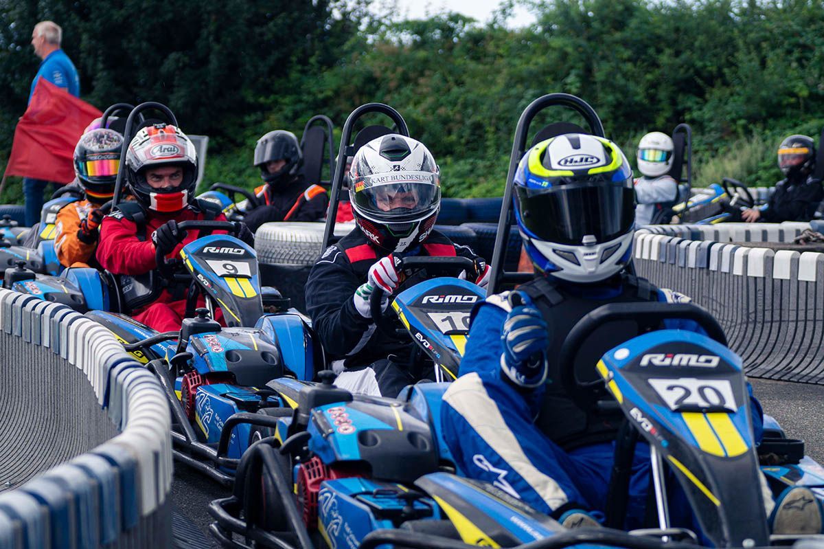 Go-Kart-Fahrer mit Helmen rasen über eine schmale Freiluftstrecke, angeführt von einem blauen Kart mit der gelben Nummer 20.