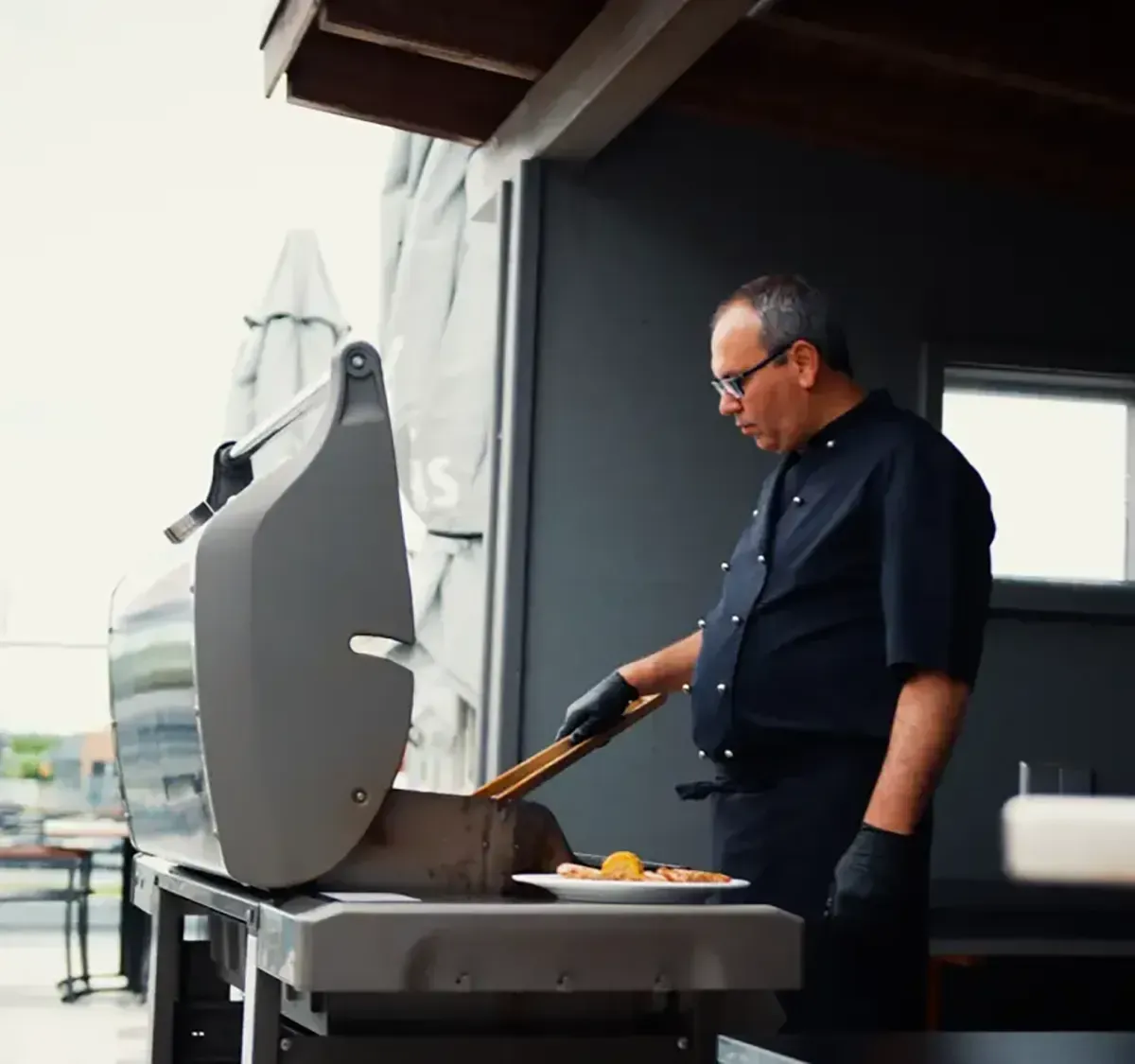 Ein Mann grillt im Freien neben einem Grill auf einer Terrasse und hält eine Grillzange über das Grillgut.