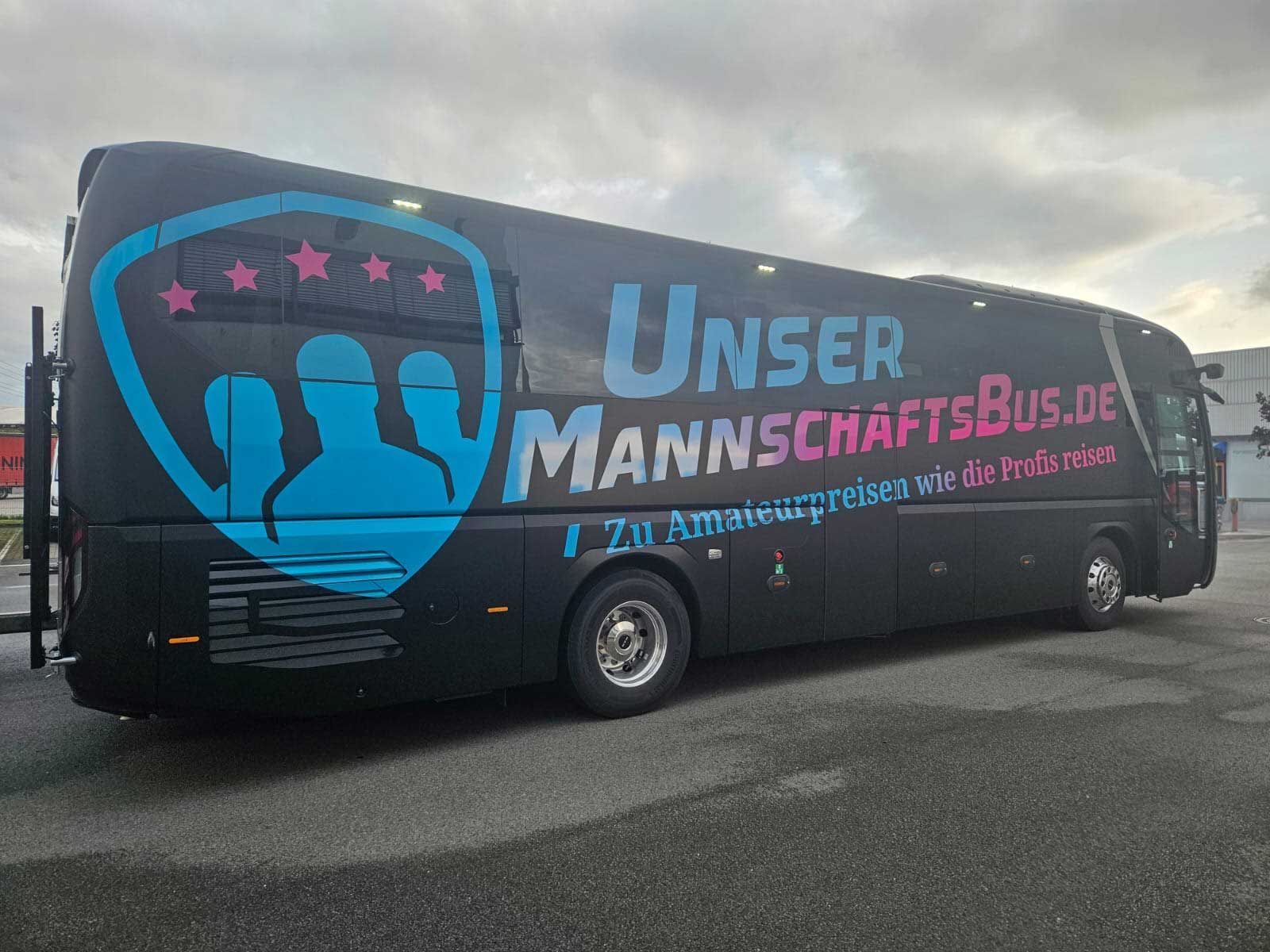 Dunkler Reisebus mit blauem Handlogo und der Aufschrift „UNSER MANNSCHAFTSBUS.de“ parkt auf dem Bürgersteig unter bewölktem Himmel