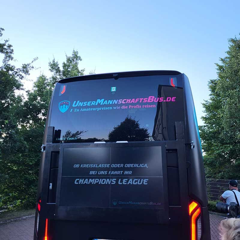 Rückansicht eines schwarzen Reisebusses auf einer von Bäumen gesäumten Straße, mit der Aufschrift „CHAMPIONS LEAGUE“ auf der Rückwand.