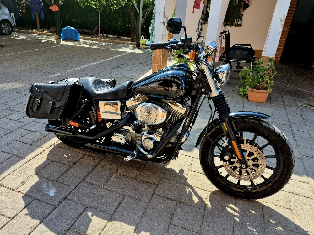 Motocicleta cruiser negra aparcada en un camino de ladrillo iluminado por el sol, junto a una casa