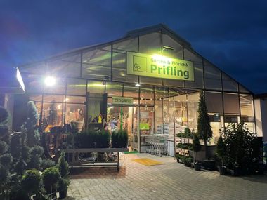 Außenansicht des Gewächshausgeschäfts in der Abenddämmerung, Schild „Garten & Floristik Prifling“ über dem Eingang, Pflanzen sind sichtbar.