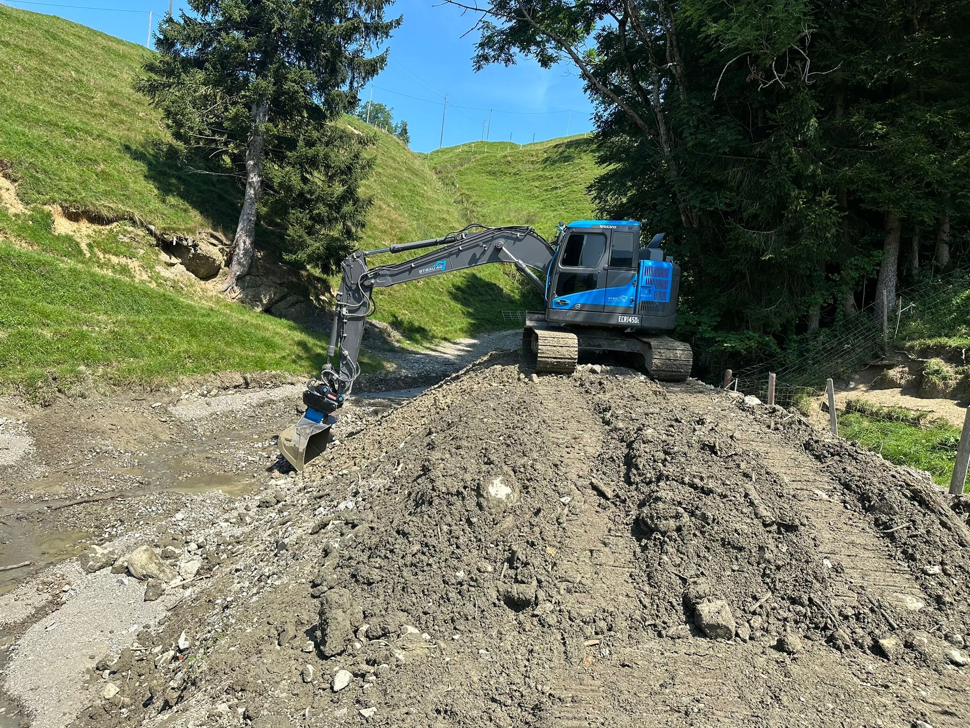 Ein blauer Bagger arbeitet an einem Feldweg.