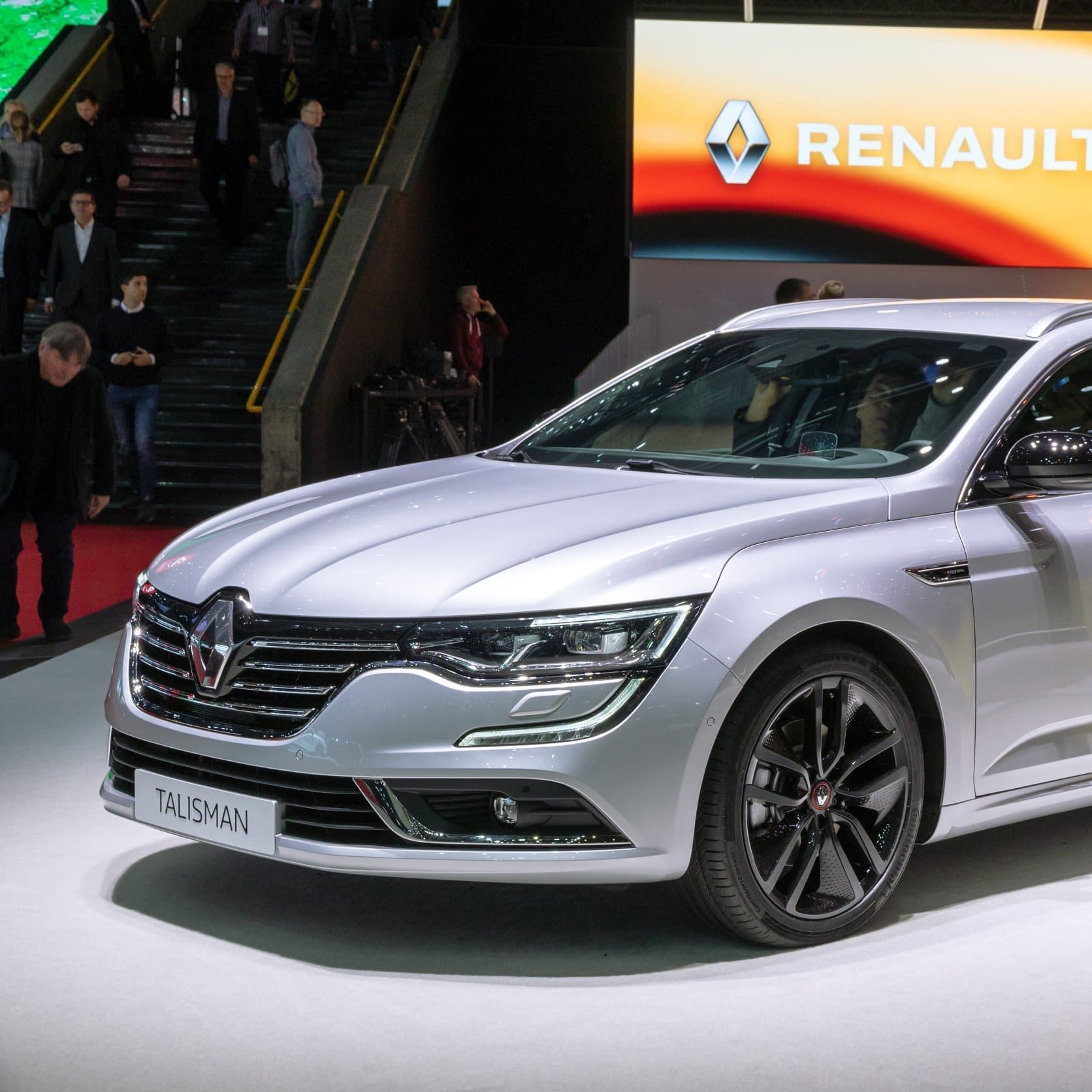Renault Talisman plateado en exhibición en un evento, con gente y un cartel de Renault al fondo.