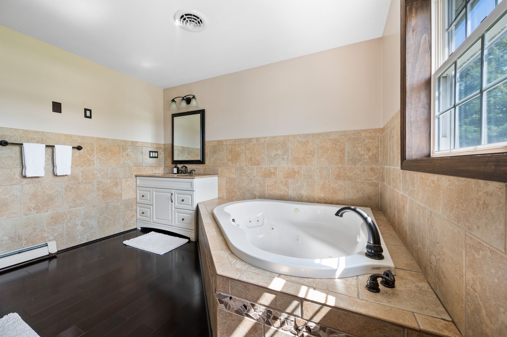 Bathroom with a jacuzzi tub, vanity, and window. Beige tile walls, dark wood floor, and white accents.