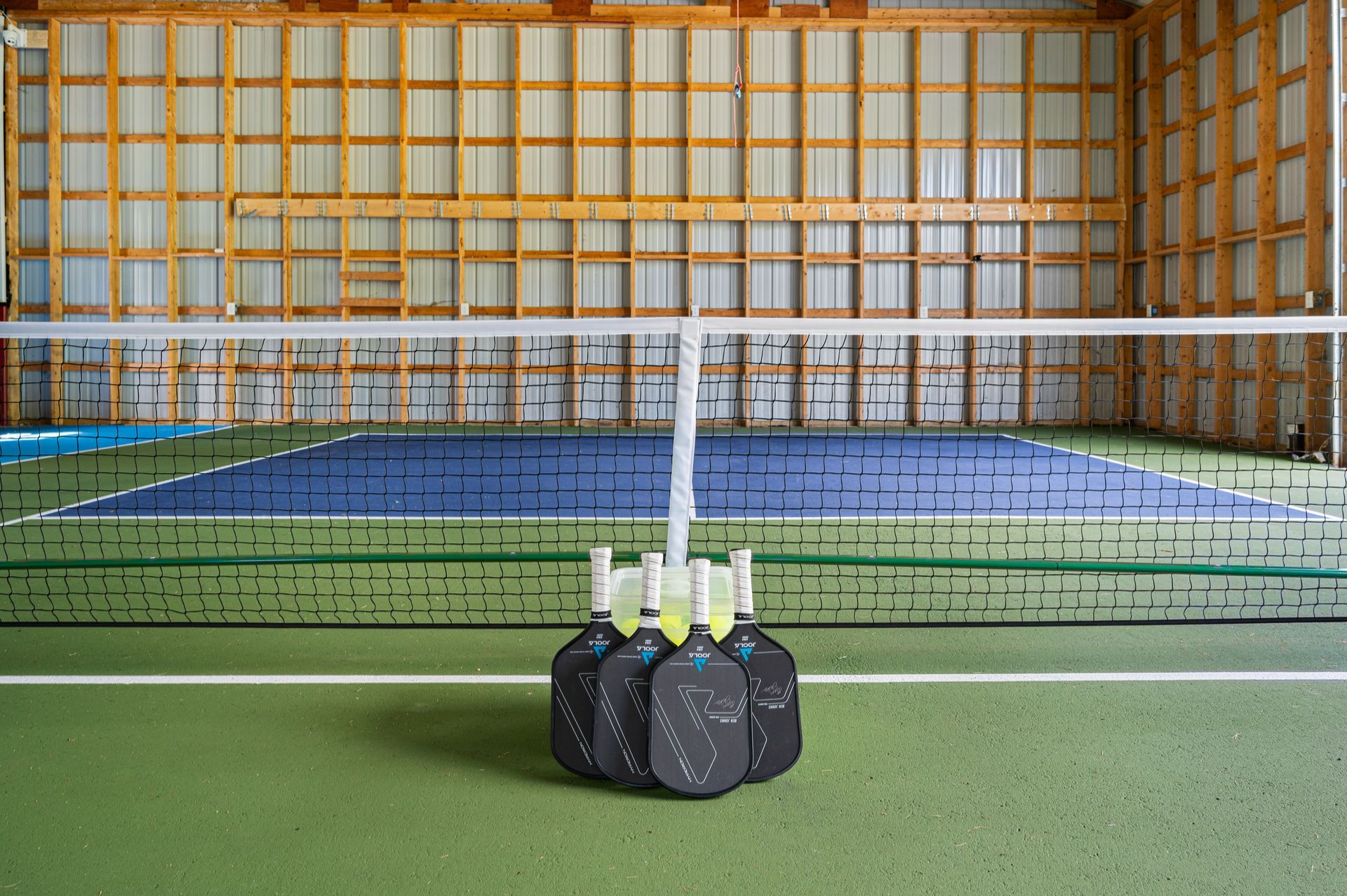 Pickleball court inside a wooden building with a net and paddles in a central holder.
